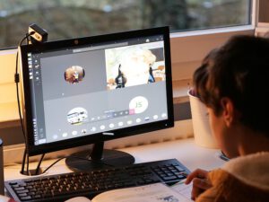 パソコン画面でオンライン学習をしている学生の後ろ姿。複数人のビデオ通話が映し出された画面と、デスク上の教材が映っている。