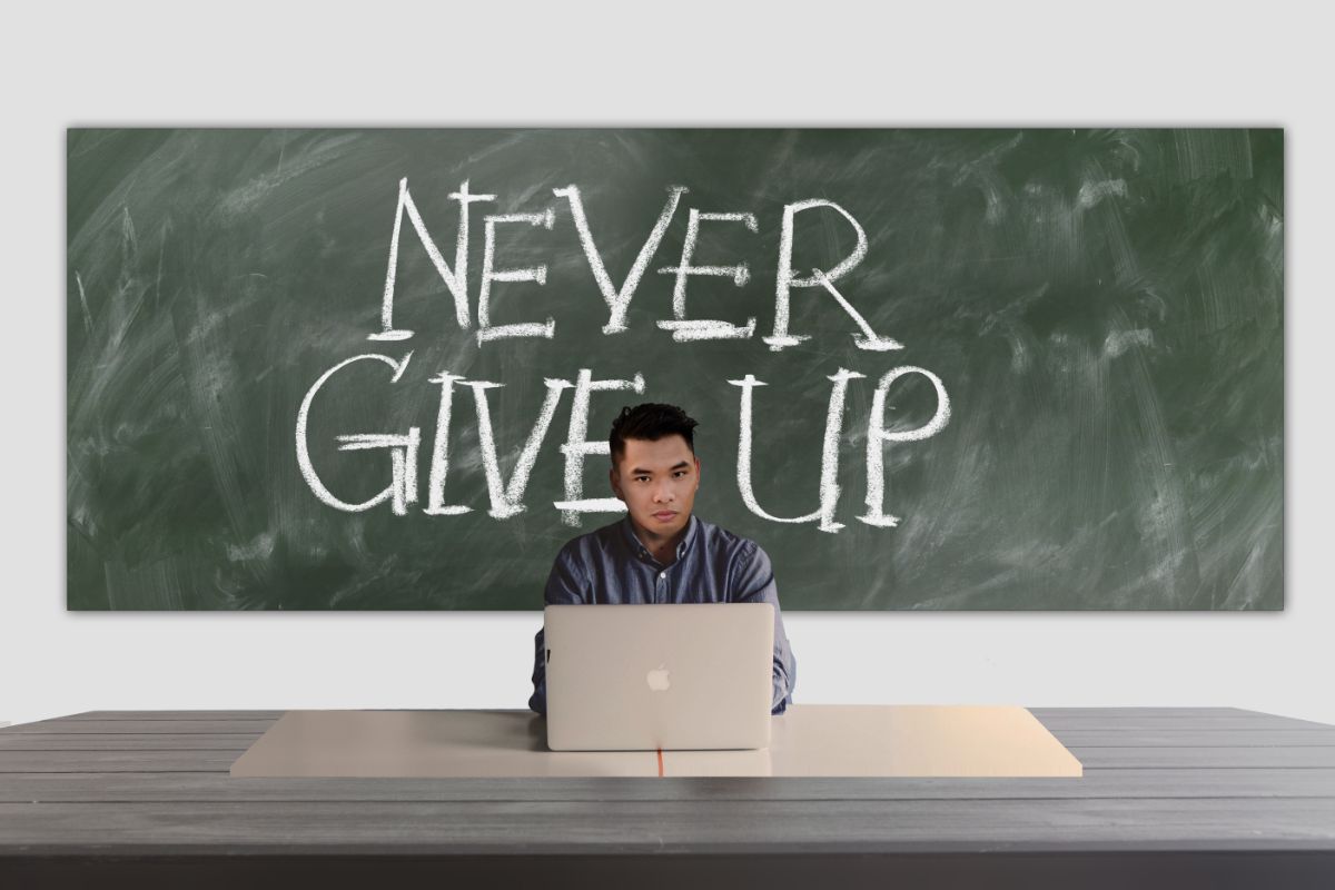 黒板に「NEVER GIVE UP」と書かれた前でノートPCに向かう男性
