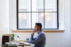 窓際のデスクでノートPCの前に座り物思いにふける男性