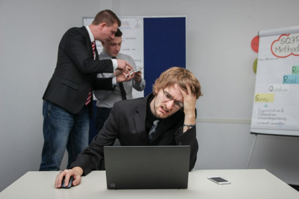 会議室でノートPCの前に頭を抱える男性｜後ろで上司が詰めている場