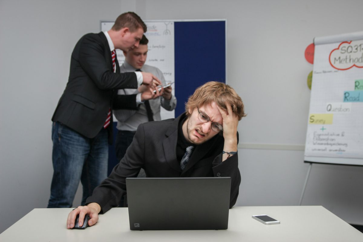 会議室でノートPCの前に頭を抱える男性｜後ろで上司が詰めている場