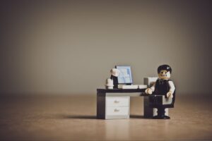 デスクでパソコンに向かい頭を抱えるレゴの人物、英文ニュースに苦戦するイメージ